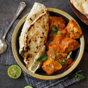 A bowl of curry with chicken pieces garnished with cilantro and accompanied by naan bread. A spoon, lime halves, and additional naan are nearby on a dark surface.