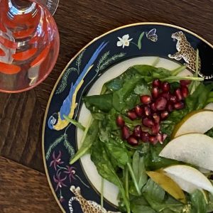 A salad with spinach, pear slices, and pomegranate seeds on a decorative plate is beside an empty wine glass with red accents on a wooden table.